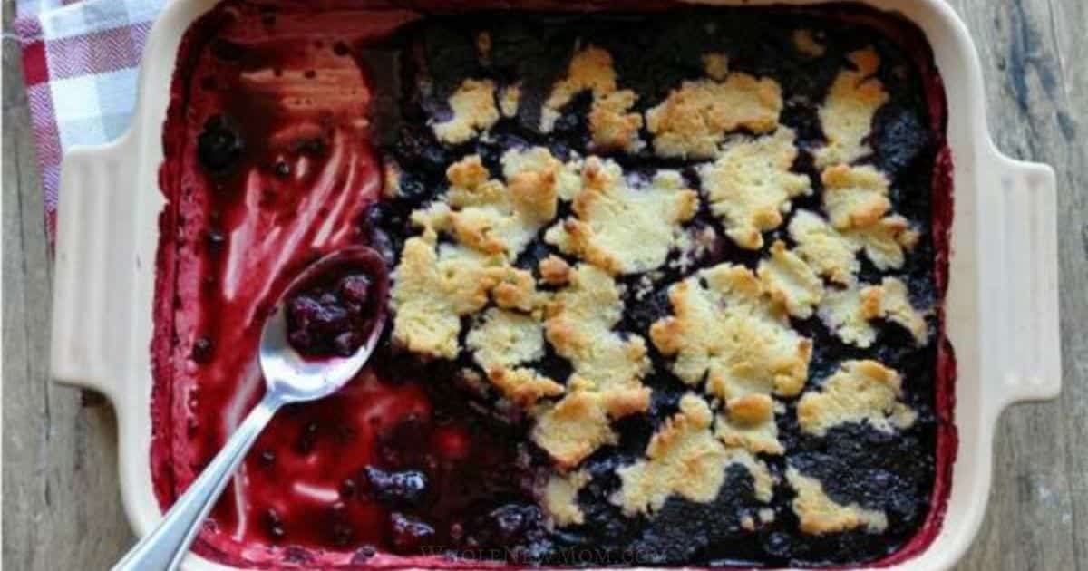 A baked berry cobbler with a golden crust, partially served, in a square dish with a spoon.