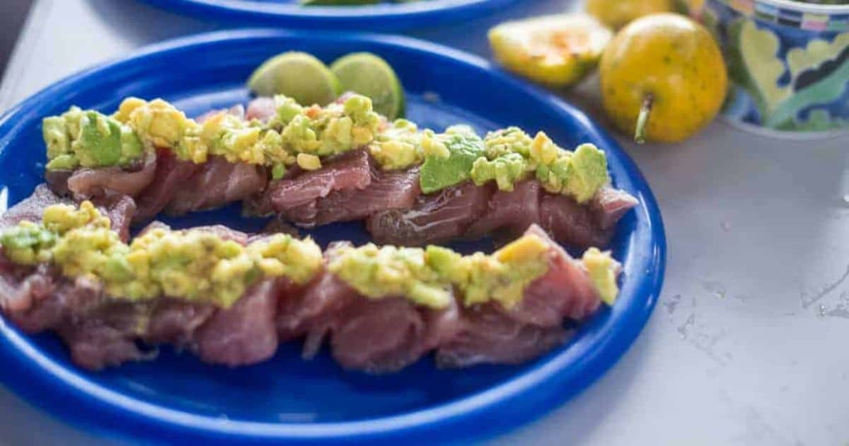 Slices of raw fish topped with avocado on a blue plate, with lime and passionfruit in the background.