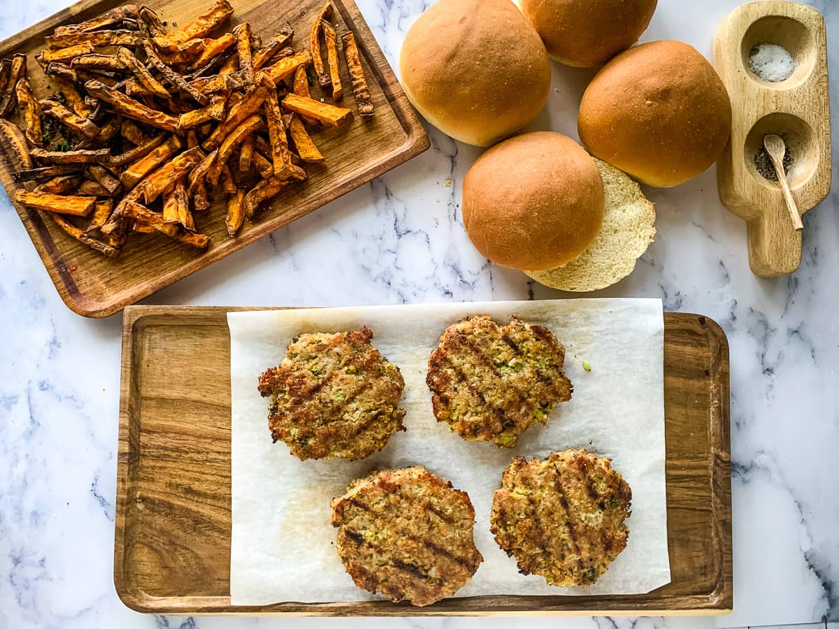 Four grilled burger patties, three buns, fries, salt, and pepper displayed on a marble and wooden surface.