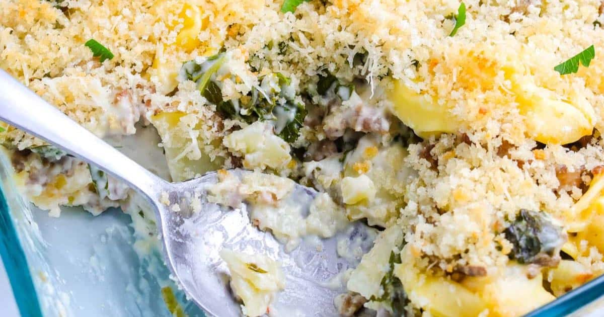 A close-up of a cheesy baked casserole with a breadcrumb topping and a serving spoon in a glass dish.