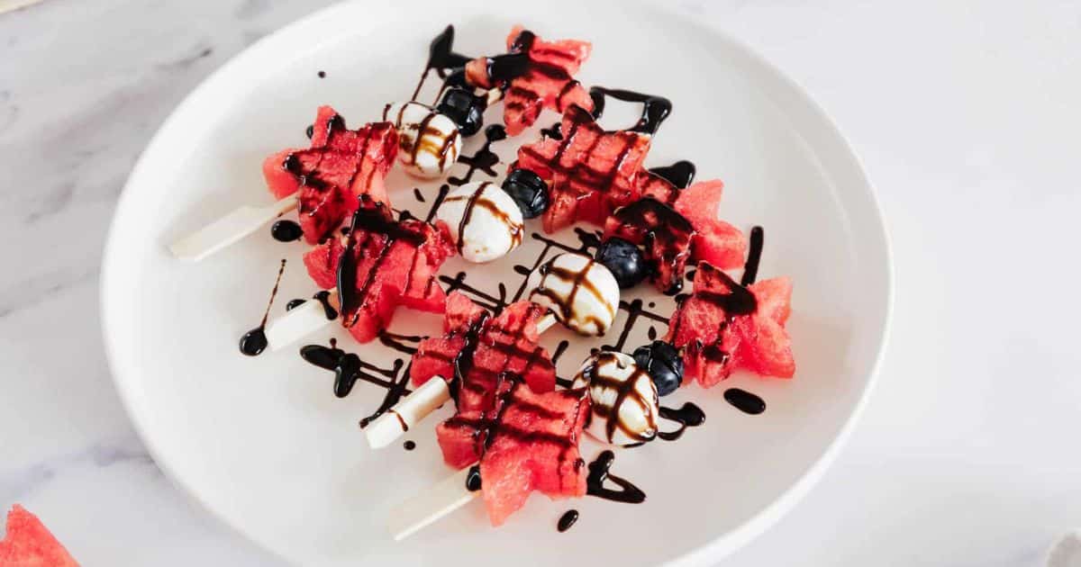 Watermelon, mozzarella, and blueberry skewers drizzled with balsamic glaze on a white plate.