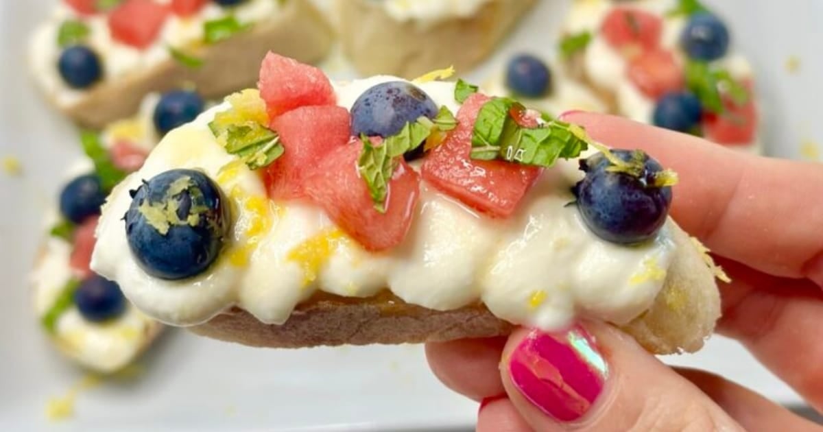 A hand holds a crostini topped with cream, blueberries, watermelon, mint, and lemon zest.