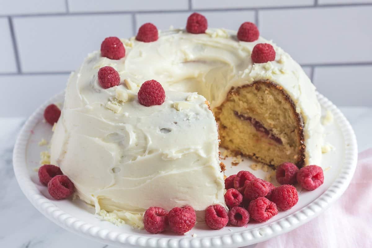 Round white frosted cake with raspberries on top and around, with one slice missing, on a white plate.