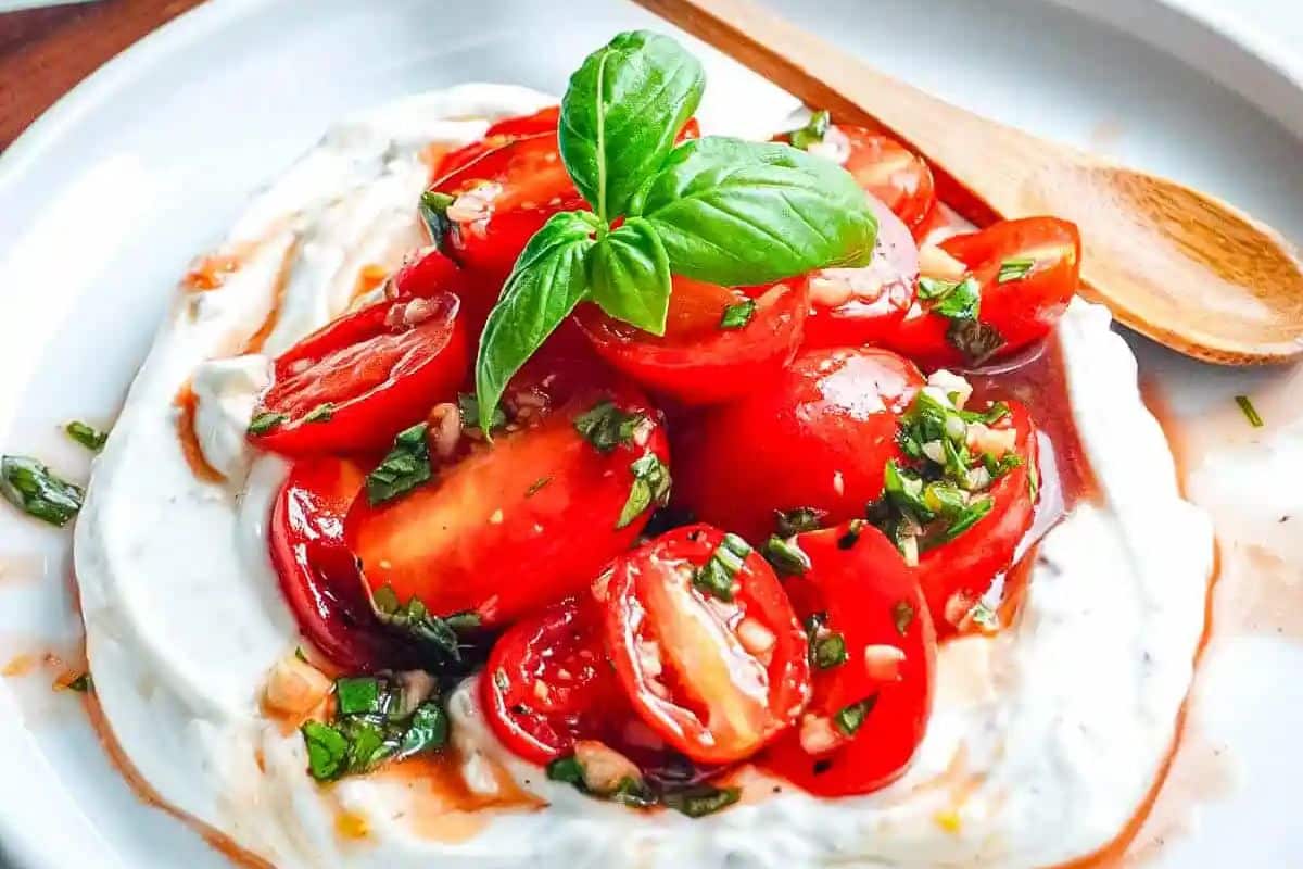 Sliced tomatoes and fresh basil on creamy yogurt, served on a plate with a wooden spoon.
