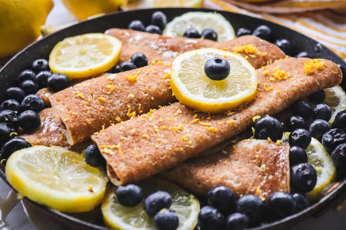 Brown crepes in a pan, topped with lemon slices, blueberries, and lemon zest.