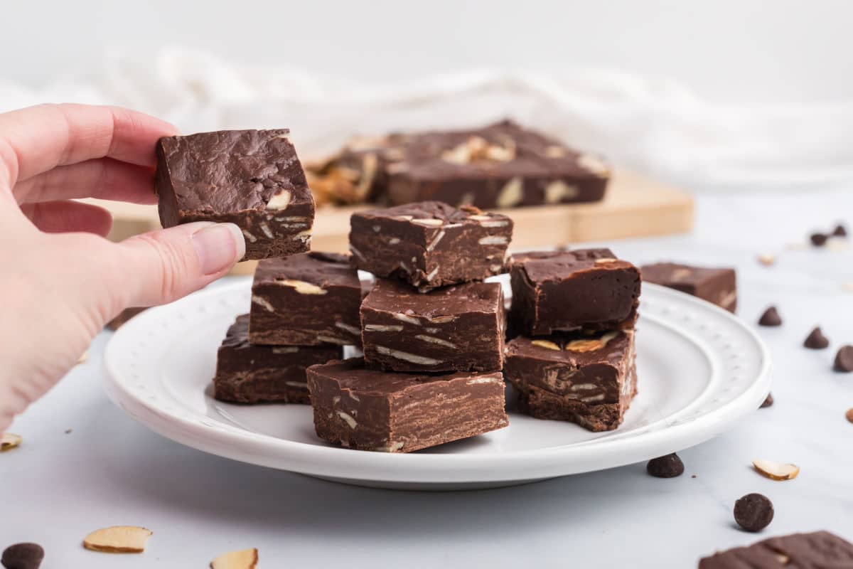 A hand holding a piece of chocolate fudge with nuts over a plate stacked with more fudge pieces.