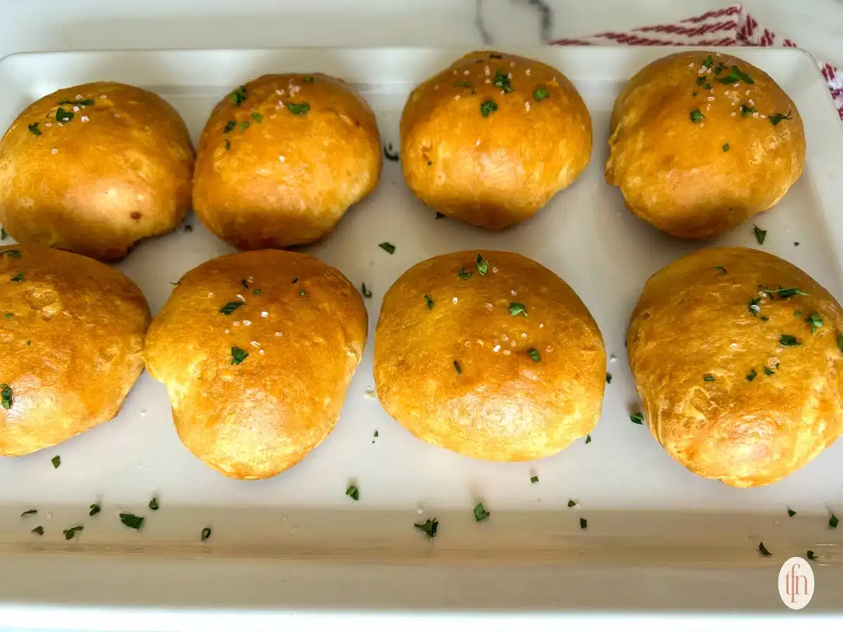 Eight golden brown bread rolls topped with herbs and salt on a white rectangular plate.