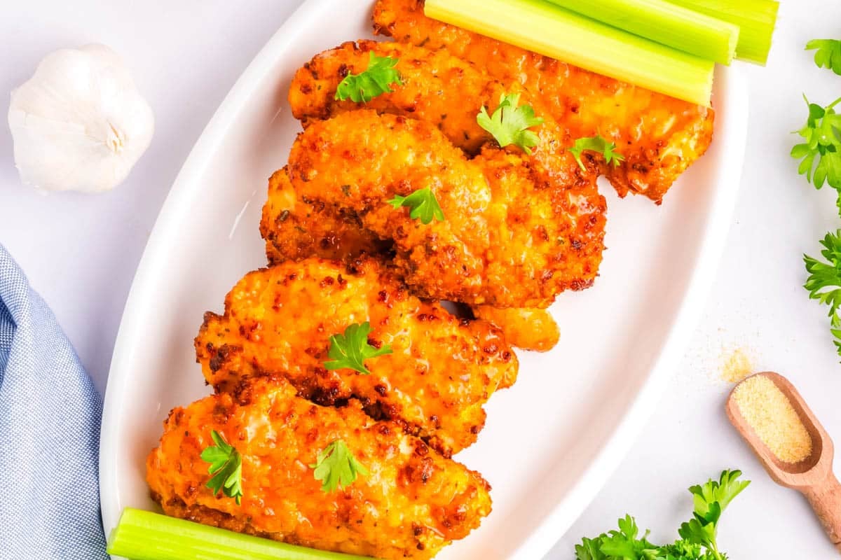 Four crispy buffalo chicken tenders garnished with parsley, served with celery sticks on a white plate.