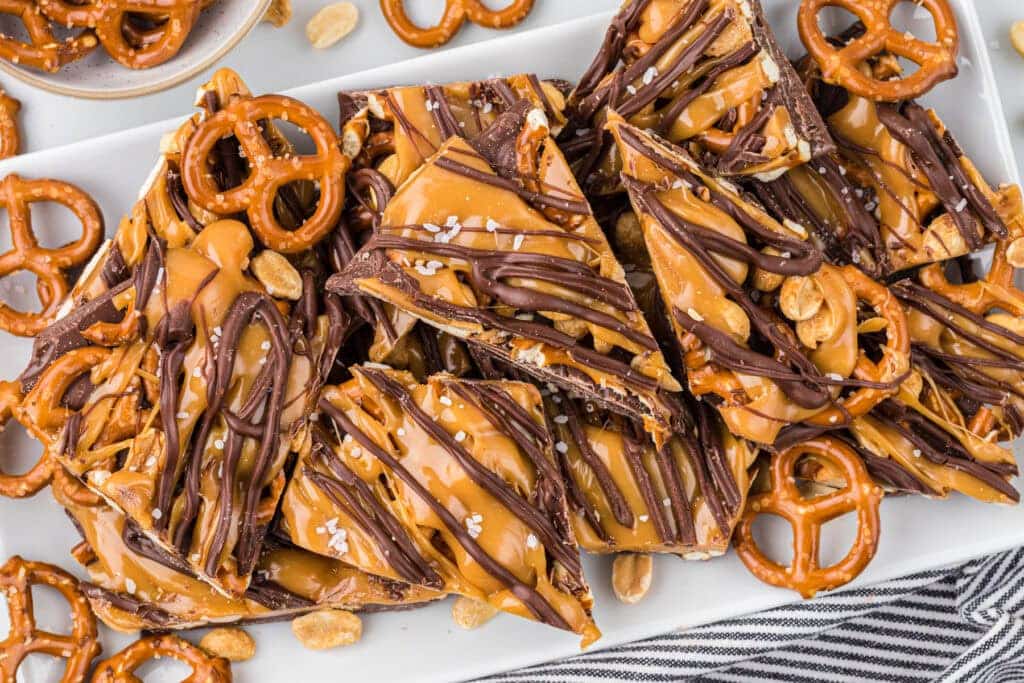 Chocolate and caramel-drizzled toffee bark with pretzels and peanuts on a white platter.