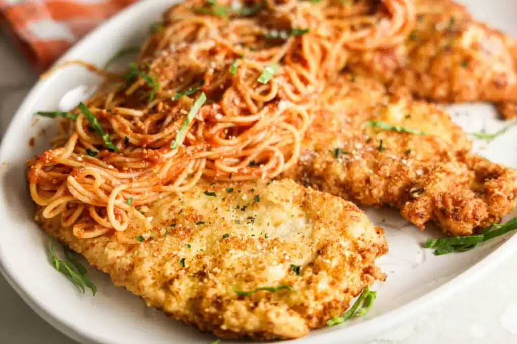 Breaded chicken cutlets served with spaghetti in tomato sauce, garnished with fresh herbs on a white plate.