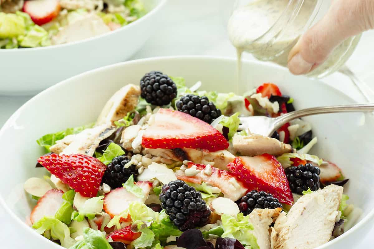 Salad with grilled chicken, strawberries, blackberries, and dressing being poured into a white bowl.