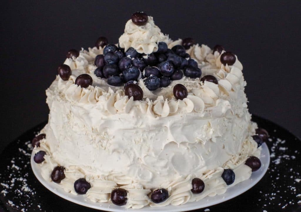 Round cake with white frosting, decorated with blueberries and grapes, on a white plate against a dark background.