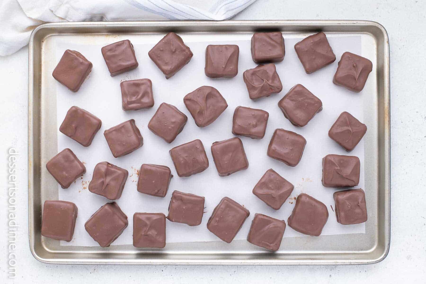 A baking sheet lined with parchment holds rows of chocolate-covered square candies.