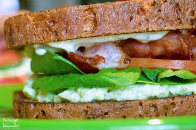 Close-up of a sandwich with whole grain bread, lettuce, bacon, tomato, and creamy spread on a green plate.