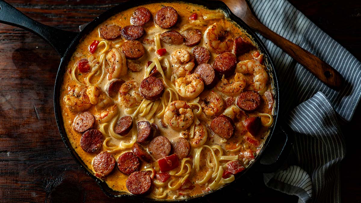 Creamy pasta in a skillet with shrimp, sausage, and red peppers, beside a wooden spoon and striped towel.