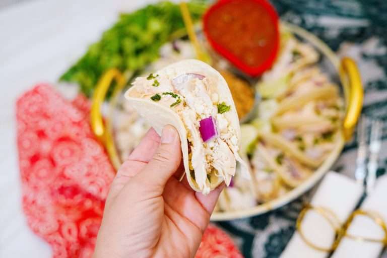 A hand holds a chicken taco with chopped onion and cilantro, with a plate of tacos and salsa in the background.