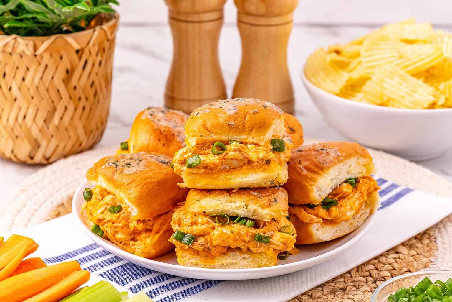 Four buffalo chicken sliders topped with green onions on a plate, with chips and vegetables nearby.