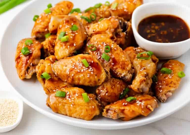 Plate of glazed chicken wings topped with sesame seeds and green onions, with dipping sauce on the side.