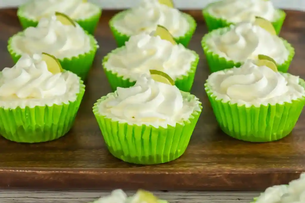 Mini cheesecakes with whipped cream in green wrappers, each topped with a small slice of lime on a wooden tray.