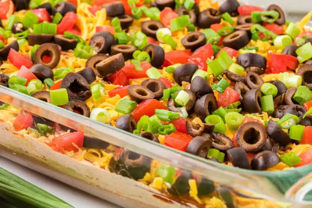 A glass dish filled with layered dip topped with chopped tomatoes, black olives, cheese, and green onions.