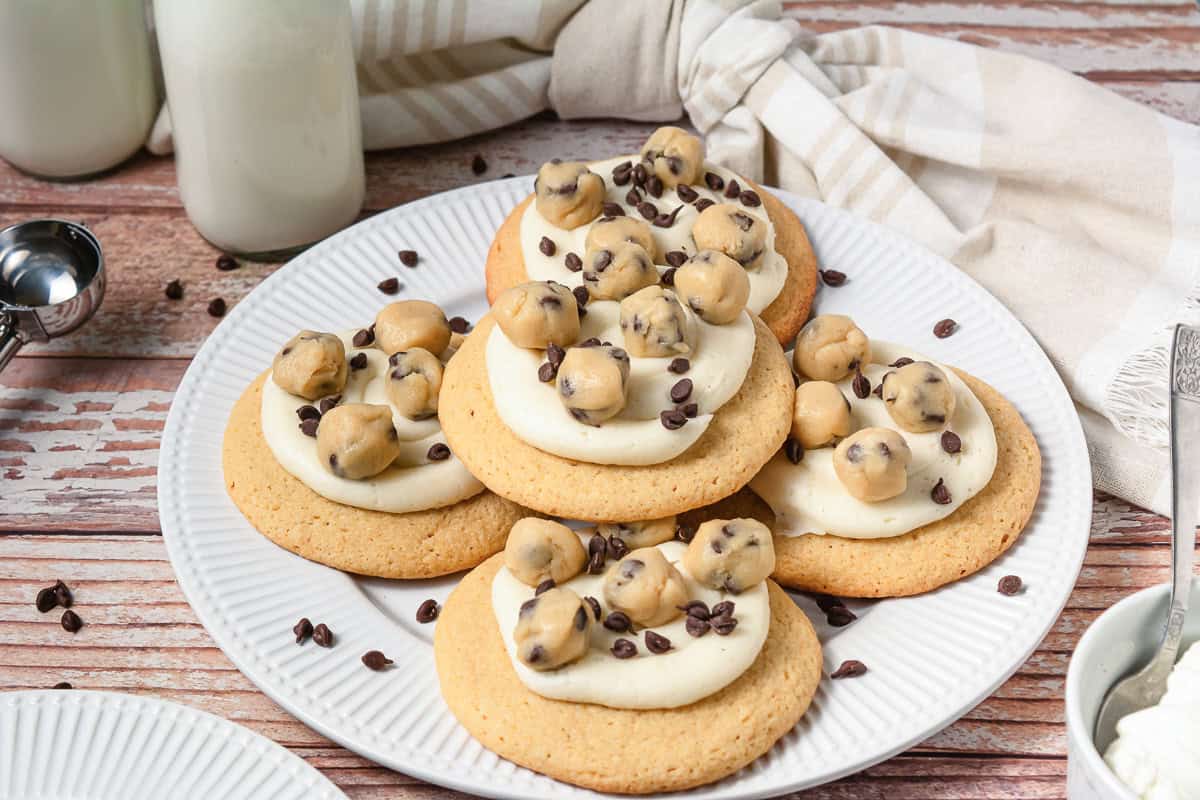 Five cookies topped with frosting, chocolate chip cookie dough bites, and chocolate chips on a white plate.