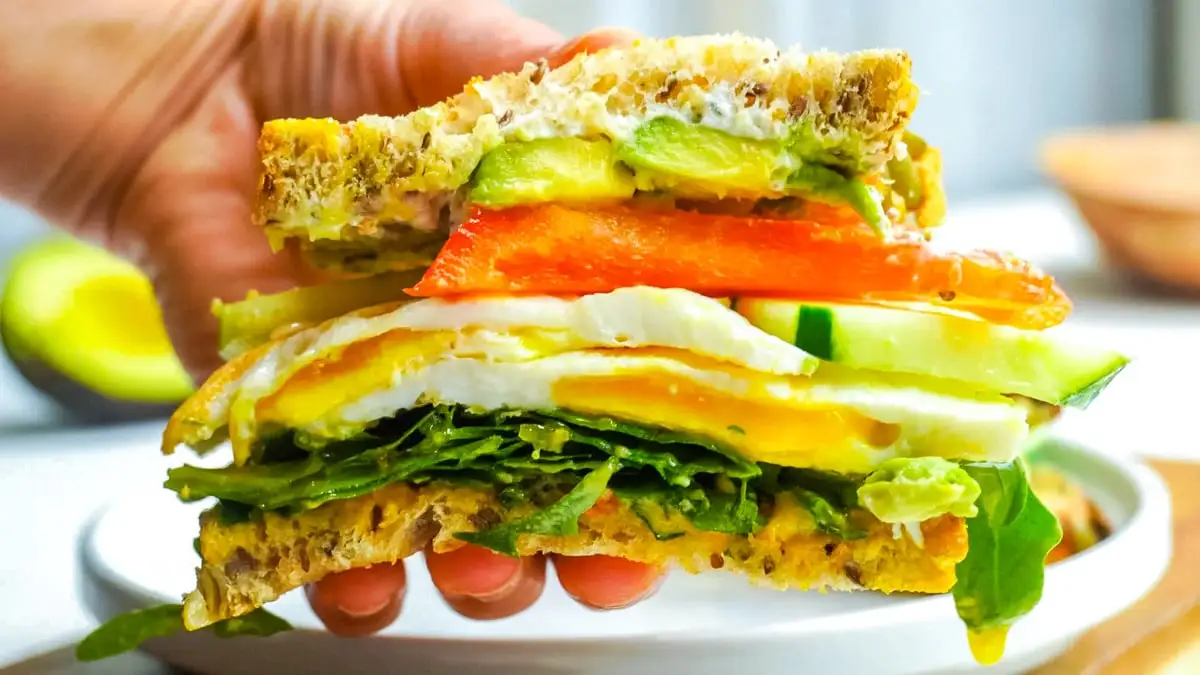 Hand holding a sandwich with egg, avocado, tomato, cucumber, greens, and multi-grain bread.