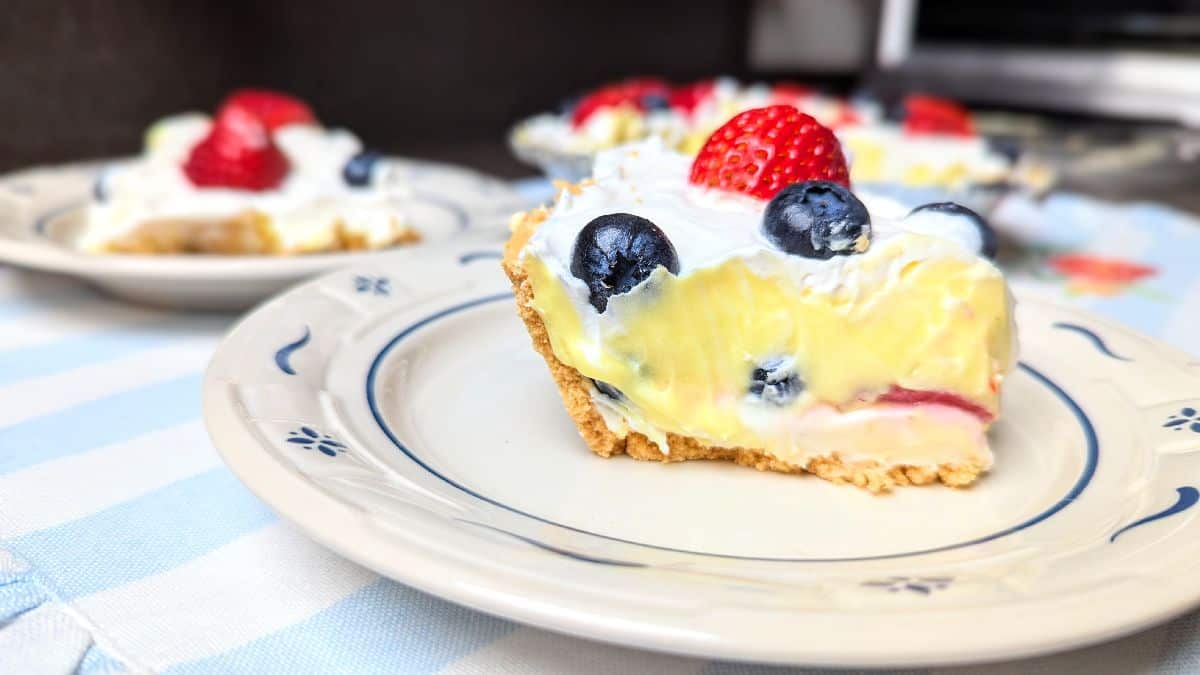 A slice of creamy pie topped with blueberries and a strawberry on a plate, with more pie slices in the background.