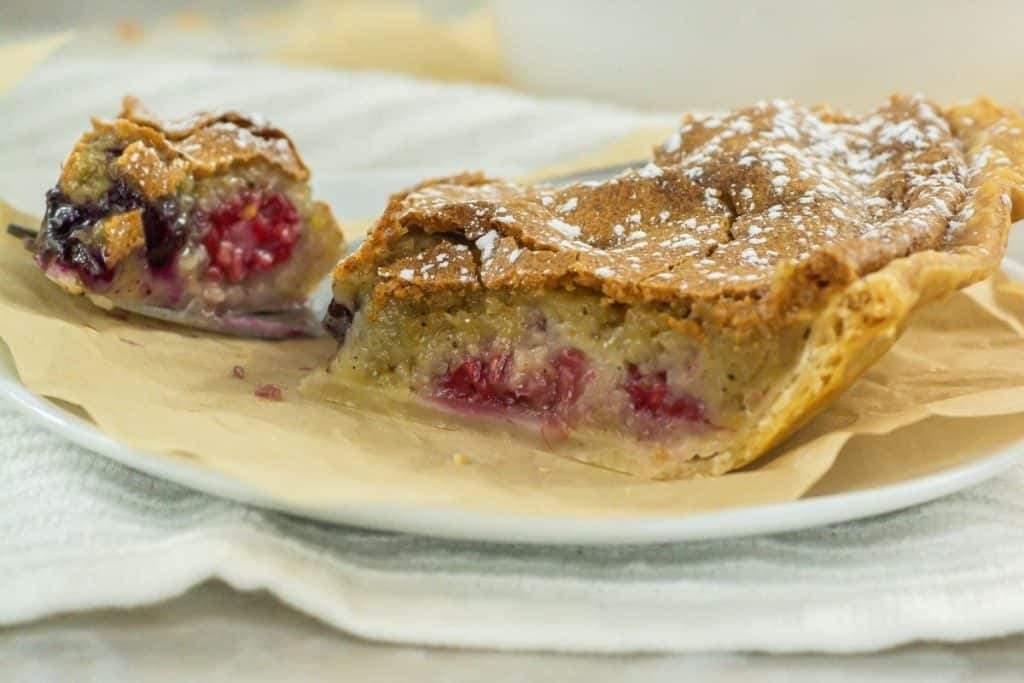 A slice of berry pie with a golden crust and powdered sugar sits next to the remaining pie on a white plate.