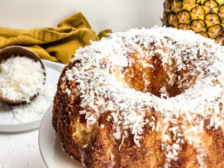 A bundt cake topped with shredded coconut, with a pineapple and a bowl of coconut flakes in the background.