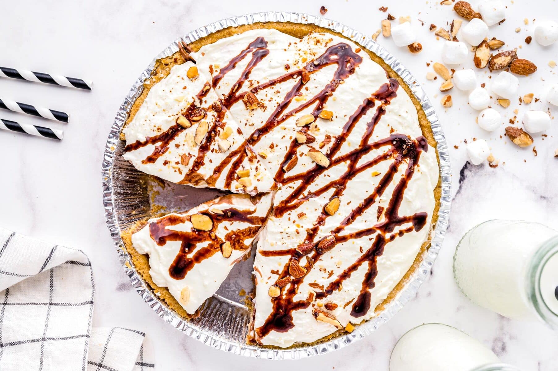 Cream pie with whipped topping, chocolate drizzle, and nuts in a foil pan, with slices cut and milk nearby.