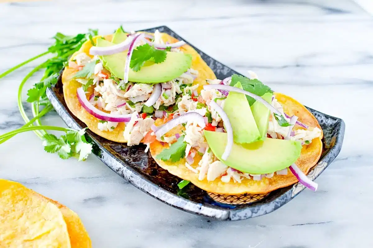 Two tostadas topped with shredded chicken, avocado slices, red onion, cilantro, and vegetables on a plate.