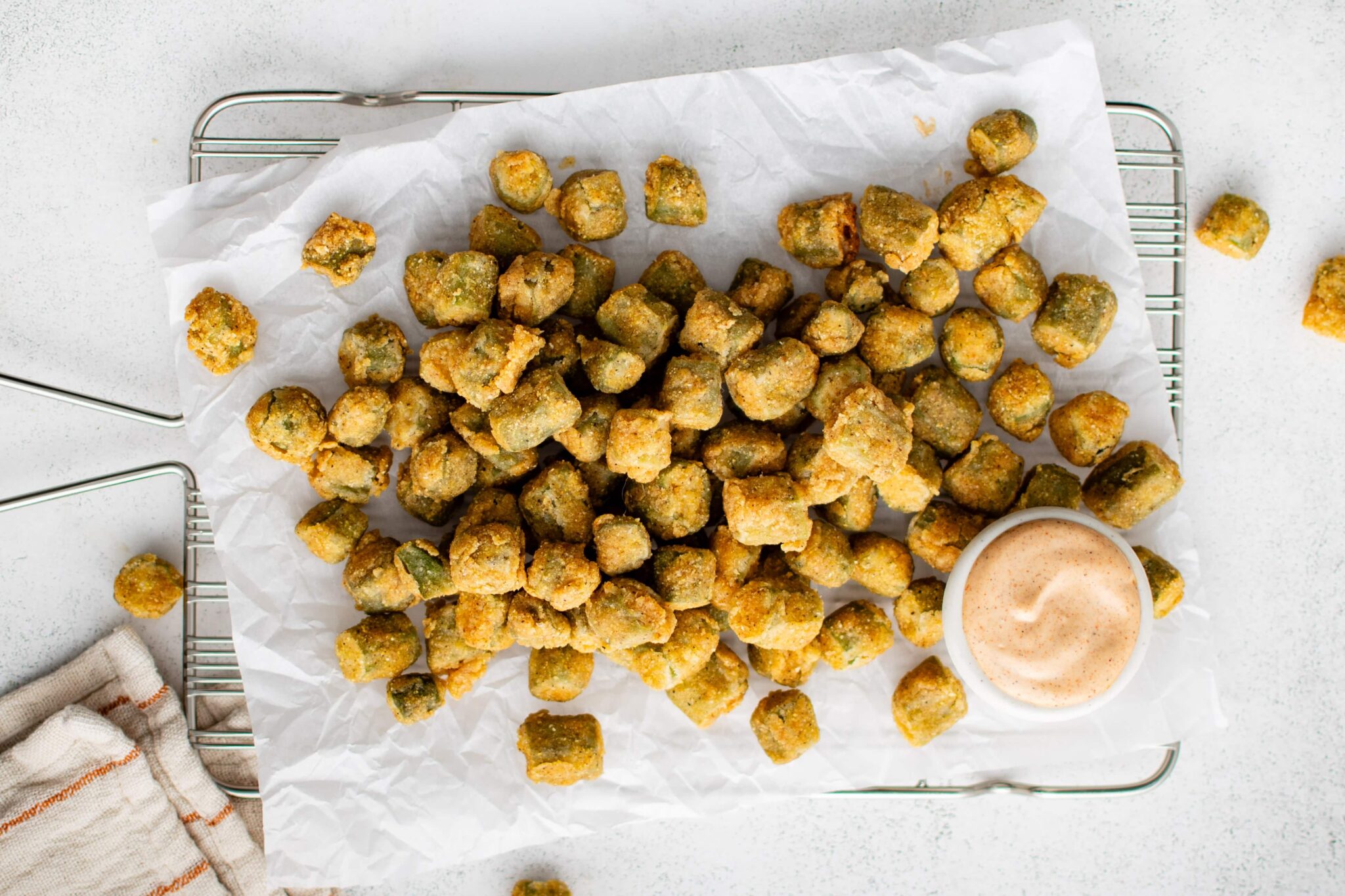 Fried okra bites on parchment paper with a small bowl of dipping sauce on a wire rack.