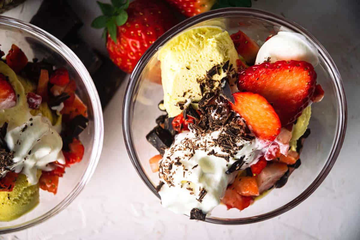 Glass bowls with yellow ice cream, whipped cream, strawberries, and chocolate shavings on a white surface.