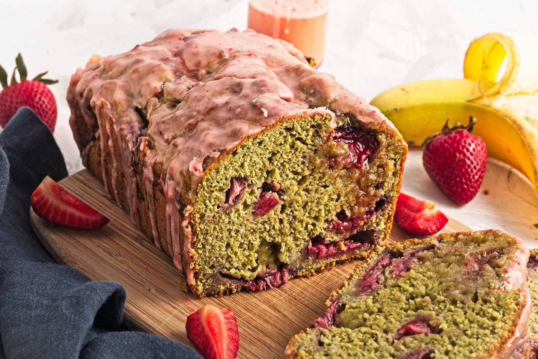 A loaf of strawberry banana bread with glaze, sliced on a wooden board, surrounded by fresh strawberries and bananas.