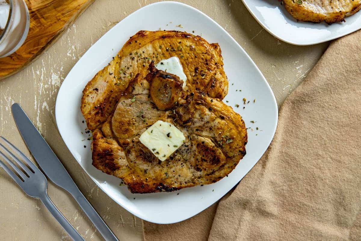 Grilled pork chops with herbs and two pats of butter served on a white plate with a fork and knife nearby.