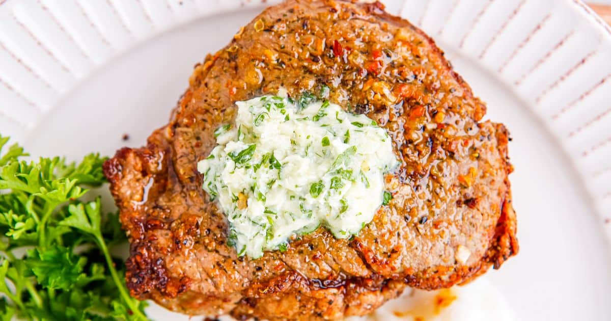 A grilled steak topped with herb butter, served on a white plate with a sprig of parsley.