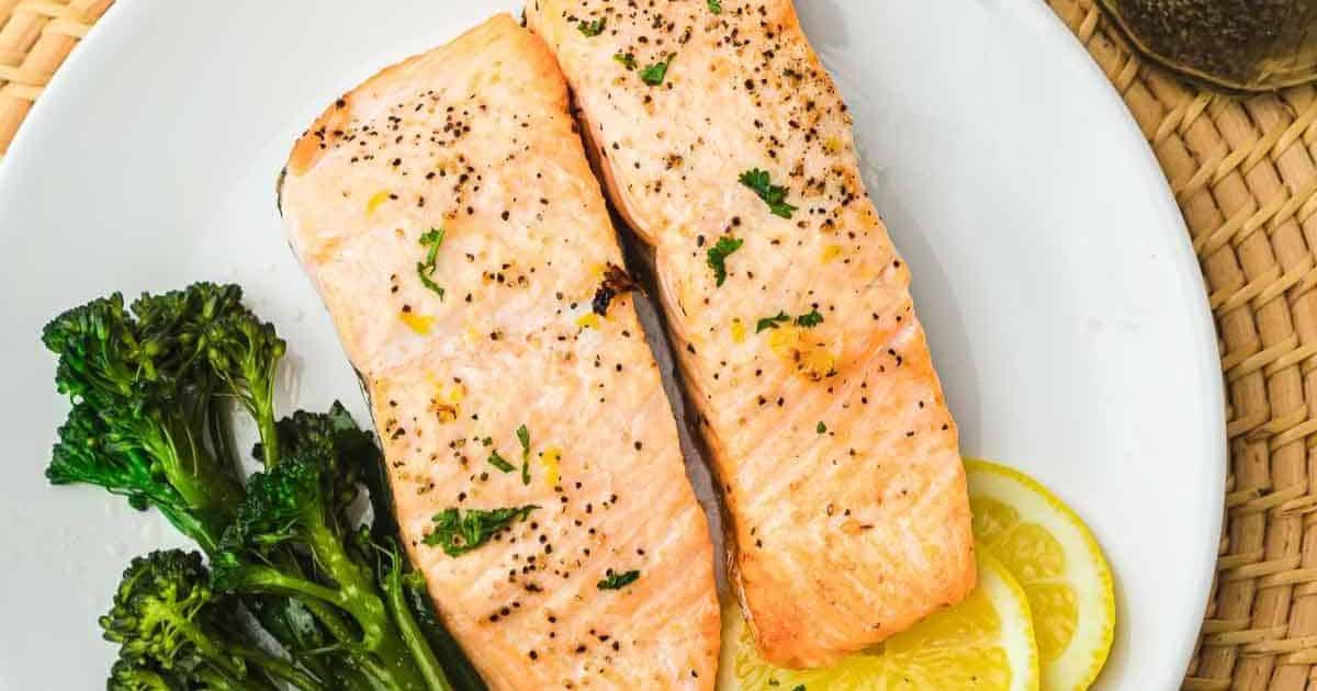 Two baked salmon fillets with pepper and herbs, served with broccolini and lemon slices on a white plate.