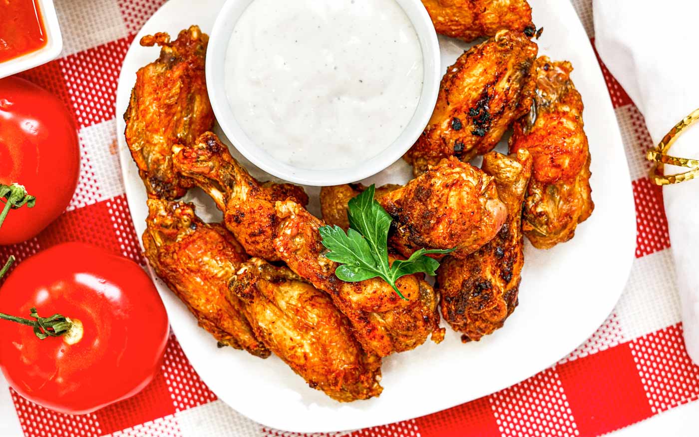Plate of spicy chicken wings with ranch dip, garnished with parsley, on a red and white checkered tablecloth.