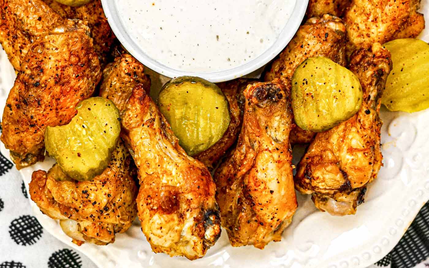Plate of seasoned chicken wings garnished with pickles, served with a cup of white dipping sauce.