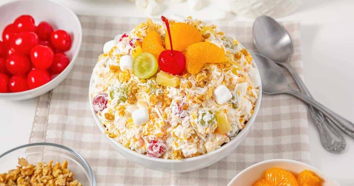 A bowl of ambrosia salad topped with fruit, marshmallows, and a cherry, next to spoons and fruit bowls.
