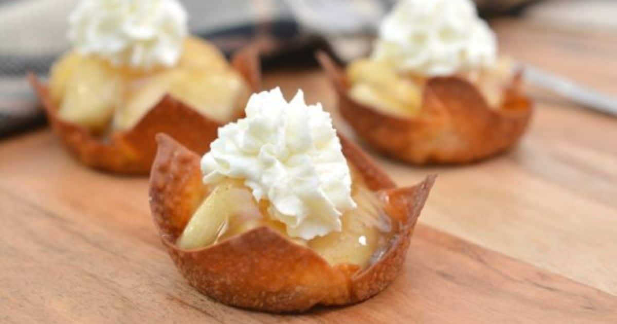 Mini apple tarts in crispy shells, topped with whipped cream, displayed on a wooden surface.
