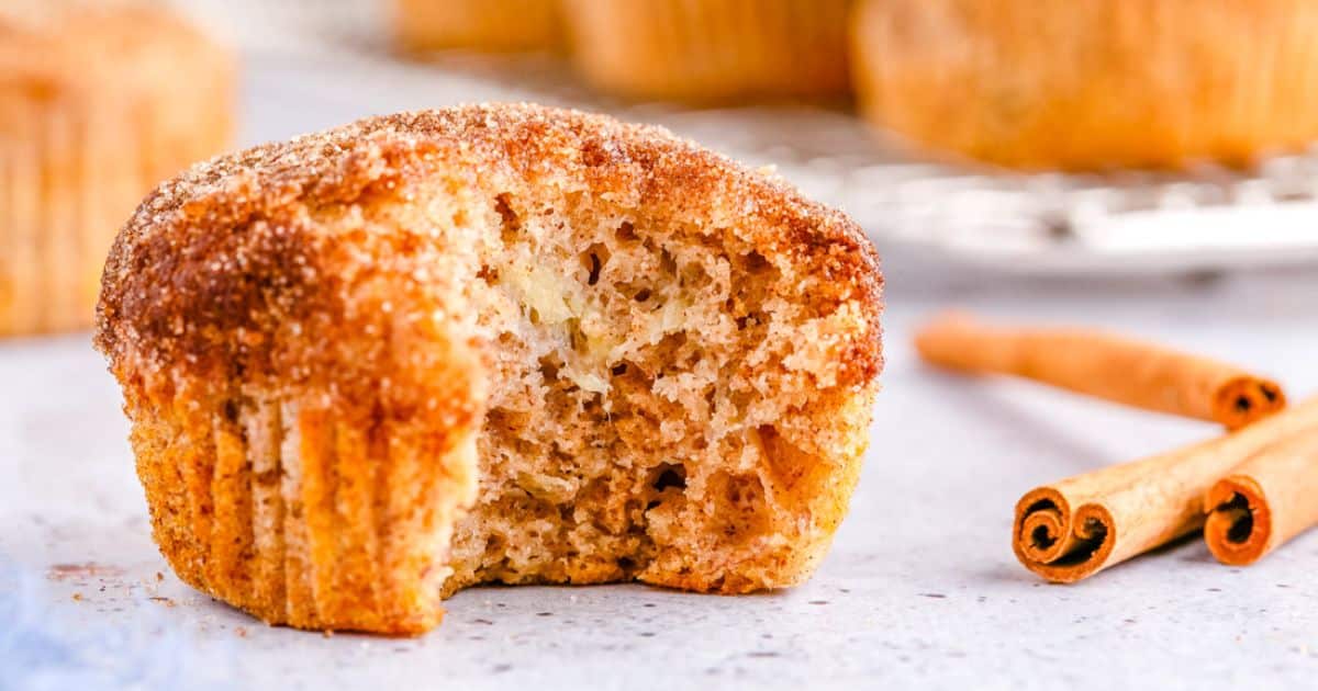 Cinnamon muffin with a bite taken out, with cinnamon sticks and more muffins in the background.