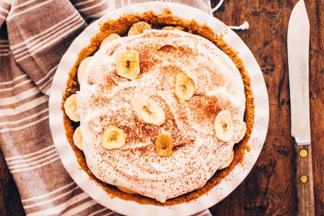 A banana cream pie with whipped topping and banana slices on a wooden table beside a striped cloth and knife.