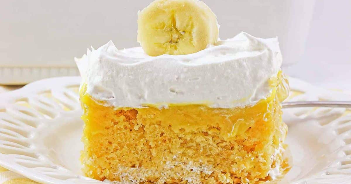 A slice of banana pudding cake with whipped topping and a banana slice on top, served on a white plate.