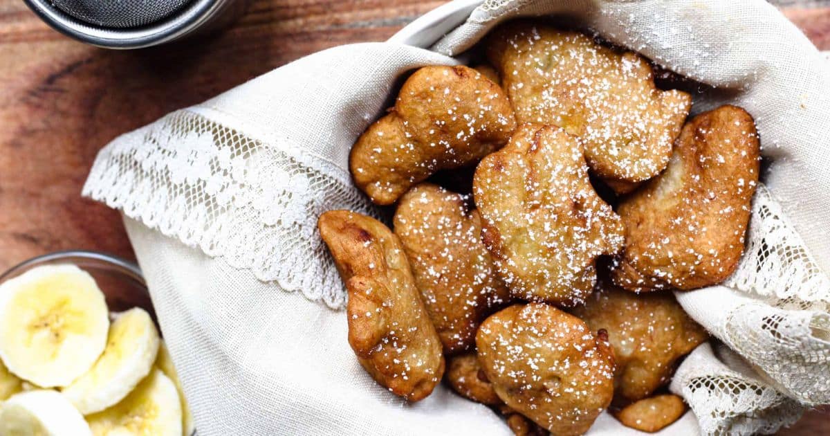 A basket of golden banana fritters dusted with powdered sugar, next to sliced bananas on a wooden table.