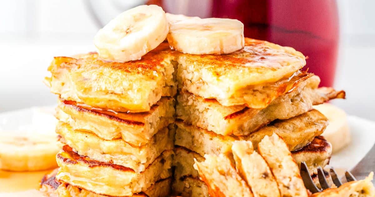 Stack of banana pancakes topped with banana slices, with syrup and a forkful taken out.