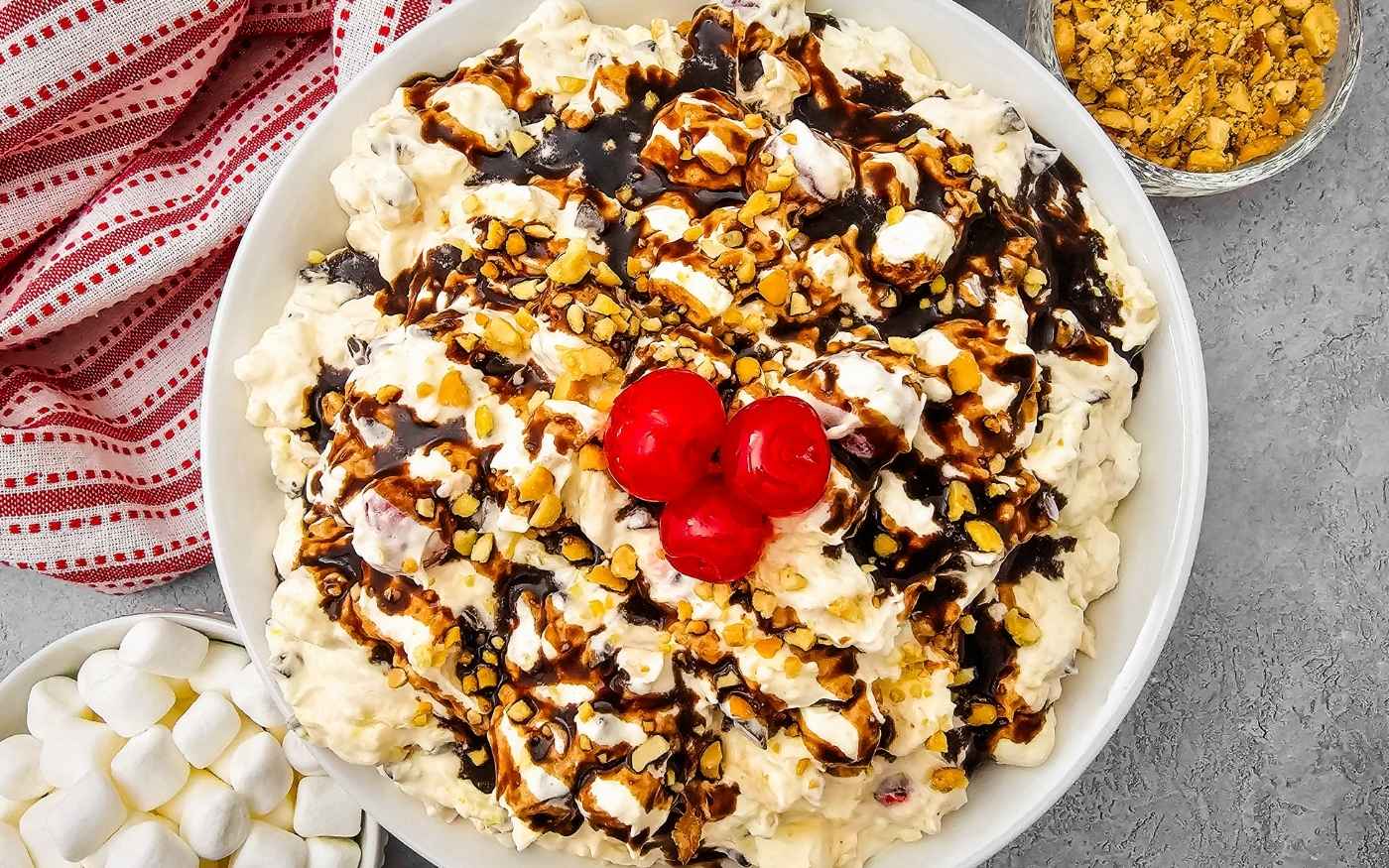 A bowl of creamy dessert topped with chocolate sauce, chopped nuts, and cherries, with marshmallows on the side.