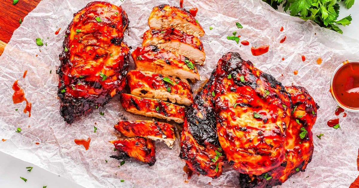 Grilled barbecue chicken breasts, sliced and whole, garnished with herbs on parchment paper with sauce on the side.