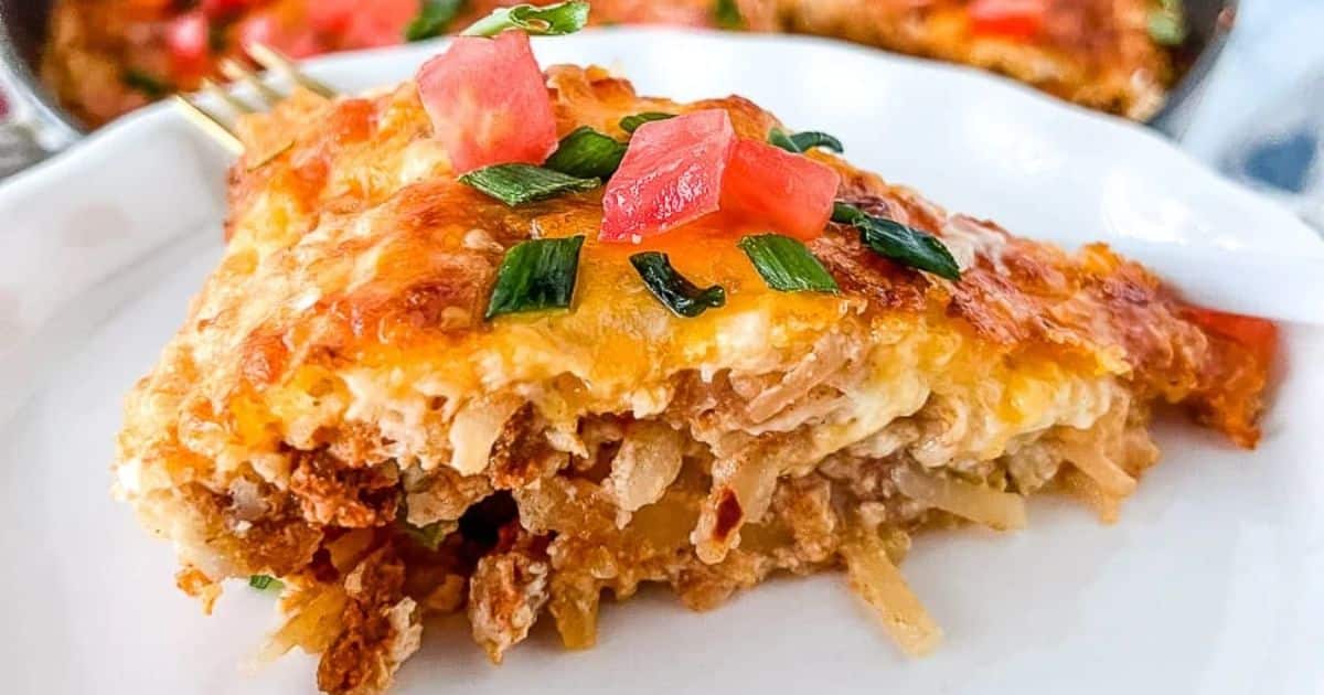 A slice of cheesy casserole topped with chopped tomatoes and green onions on a white plate.