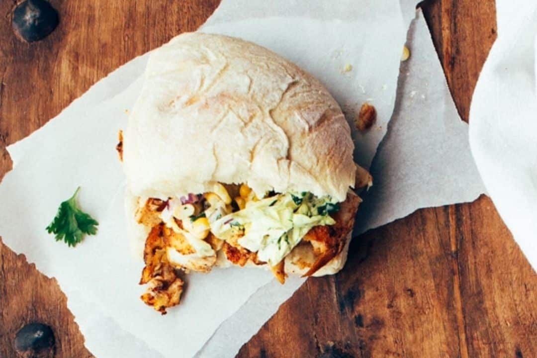 Chicken sandwich with coleslaw filling on parchment paper, partially eaten, on a wooden table.
